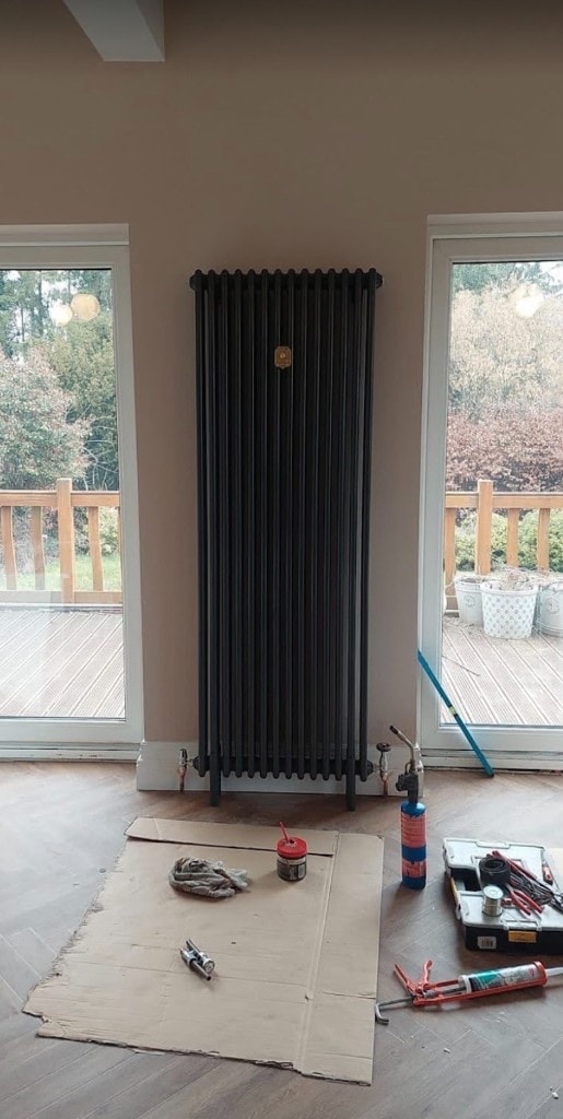 Black vertical column radiator newly installed between patio doors with plumbing tools on the floor